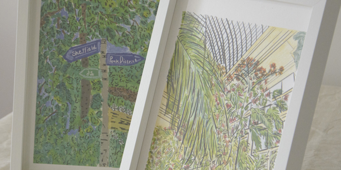 Two framed art prints displayed on a light fabric surface. The left print features a green leafy scene with a signpost pointing to “Sheffield” and “Peak District.” The right print shows a botanical illustration of tall green plants under a glass roof structure, with soft yellow and pink tones in the background.