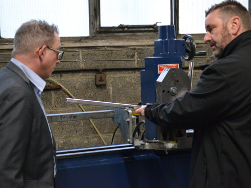 Two people standing next to a metalworking machine inside an industrial workshop. One person is holding a long metal rod positioned in the machine, while the other observes. The background shows a concrete wall, windows, and electrical wiring.