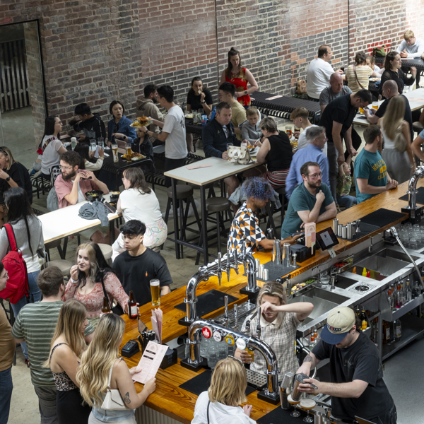 Inside the Cambridge Street Collective food hall there are rows of long tables filled with people eating and chatting. To one side is a bar where people are being served drinks.