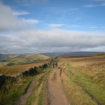 A rough track winding through hills in the countryside. In the distance you can see various groups of people out walking.