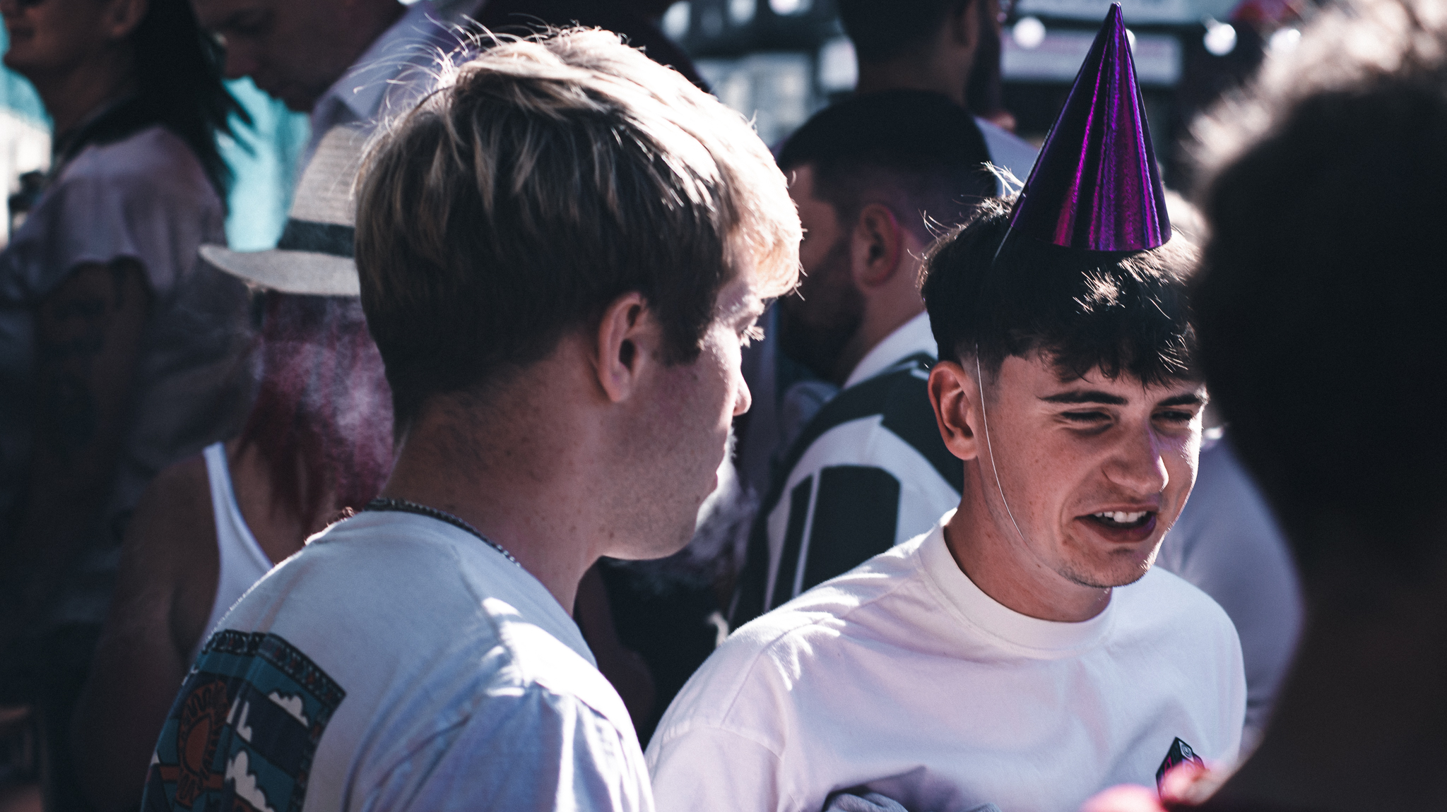 Two people talking in a crowd at an outdoor event, with one wearing a shiny purple party hat as others gather around in the sunlight.