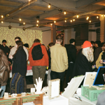 Busy indoor market event with visitors browsing stalls inside a large warehouse-style venue. String lights hang from the ceiling, and a wall of warm fairy lights glows in the background. In the foreground, tables display handmade artworks, prints and decorative items, including sculpted hands and framed pieces. Attendees in coats and colourful hats gather around stalls, creating a lively atmosphere typical of a craft market or community arts event.