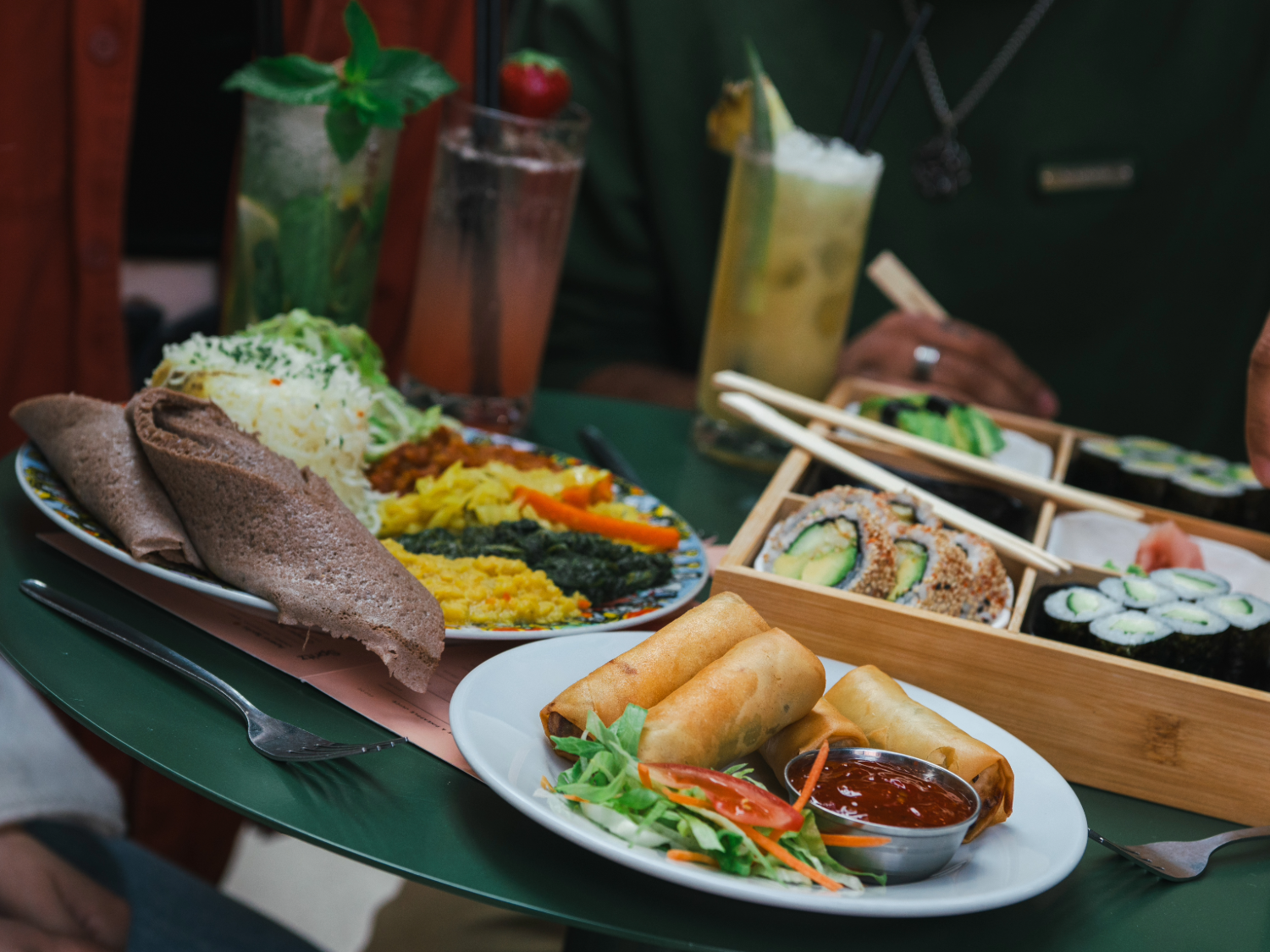 Close-up of a green table filled with assorted dishes and drinks. A plate of golden spring rolls with dipping sauce and salad sits in the foreground. Behind it, a colourful platter features flatbread, shredded cabbage, and vibrant vegetable sides. A wooden tray holds sushi rolls, nigiri, and pickled ginger with chopsticks resting on top. Three tall glasses of cocktails garnished with fruit and herbs are also visible, creating a fresh and vibrant dining scene.
