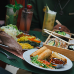 Close-up of a green table filled with assorted dishes and drinks. A plate of golden spring rolls with dipping sauce and salad sits in the foreground. Behind it, a colourful platter features flatbread, shredded cabbage, and vibrant vegetable sides. A wooden tray holds sushi rolls, nigiri, and pickled ginger with chopsticks resting on top. Three tall glasses of cocktails garnished with fruit and herbs are also visible, creating a fresh and vibrant dining scene.