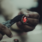 Close-up of artisan hands using pliers to craft a pendant with a deep red gemstone in a decorative metal setting, surrounded by similar pieces on a workbench.