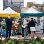 A lively outdoor market scene with people gathered around stalls covered by yellow and green canopies. Various individuals are browsing items, chatting, and holding bags or flowers. Display boards and tables with informational materials are set up under the tents. The background shows older brick buildings and a tall modern structure, while the foreground features plants and a paved area.