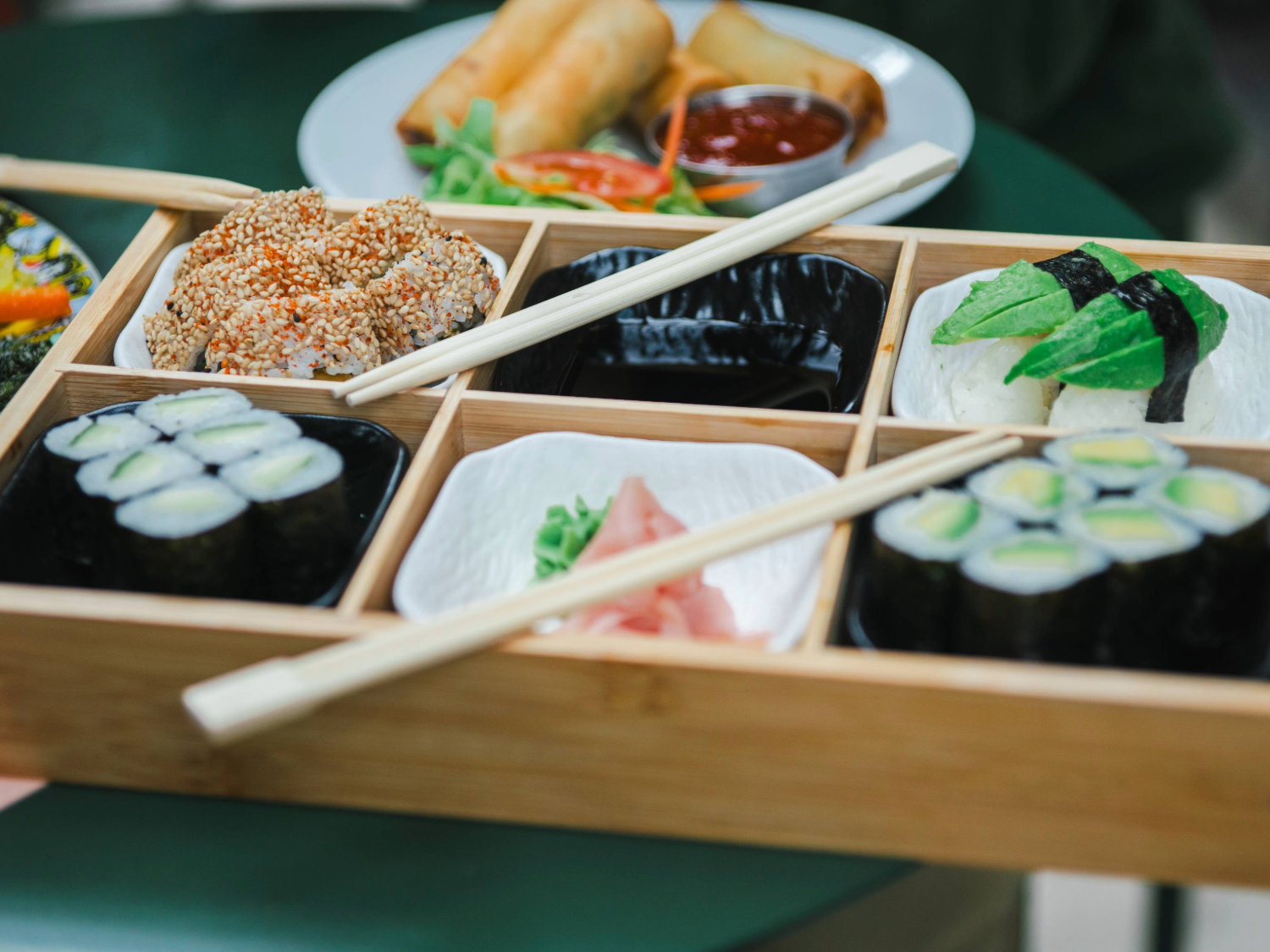 A wooden bento-style tray filled with assorted sushi, including maki rolls, nigiri topped with avocado, and sesame-coated pieces. Two pairs of chopsticks rest across the tray. In the centre, a small dish holds pickled ginger and wasabi. Behind the tray, a plate with golden spring rolls and a dipping sauce is partially visible. The food is arranged on a green table, creating a fresh and vibrant presentation.