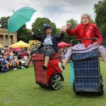 Two performers dressed in vintage-style clothing ride motorised shopping trolleys covered in tartan fabric during an outdoor event. One holds a bright green umbrella while both raise their arms in animated gestures. Behind them, a crowd watches near colourful market-style tents set up on a grassy area with trees and a large classical building in the background.