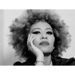 Black-and-white portrait photograph of a person with voluminous curly hair, resting one side of the face on a hand. The image is closely cropped, focusing on the face, hair and upper shoulders. The subject wears earrings and a bracelet, with defined eye makeup visible. A plain, softly lit background keeps attention on the facial features and pose.