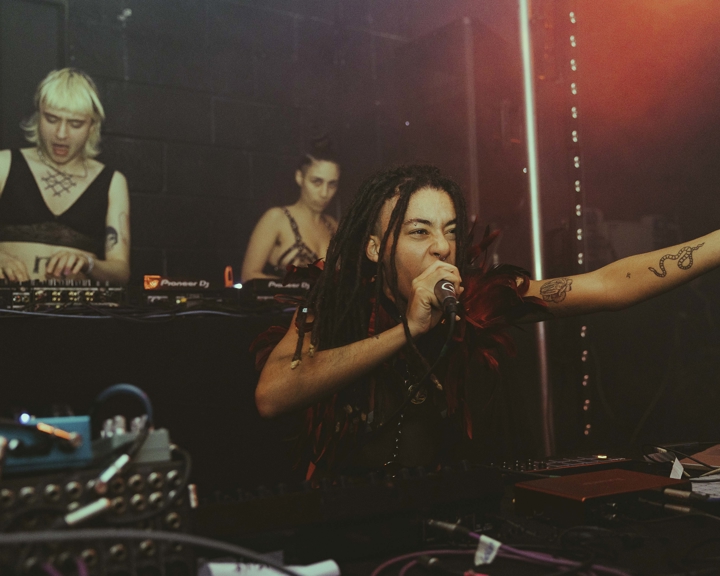 Three performers on stage at No Bounds festival. One is singing into a microphone while the other two are playing records on turntables and using a mixer.