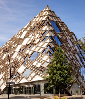 The exterior of the Diamond Building in Sheffield. Striking modern building with a distinctive diamond-shaped design featuring a geometric lattice of wooden beams and large glass panels. The structure has a pyramid-like form with multiple triangular windows, creating a bold architectural statement. Surrounding the building are trees, a paved street, and older brick buildings under a clear blue sky.