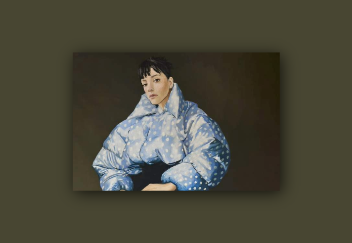 A person wearing an oversized, light blue puffer jacket with white polka dots. The jacket has exaggerated, rounded sleeves and a high collar, creating a bold, sculptural silhouette. The background is a plain, dark neutral tone, emphasising the striking design of the garment.