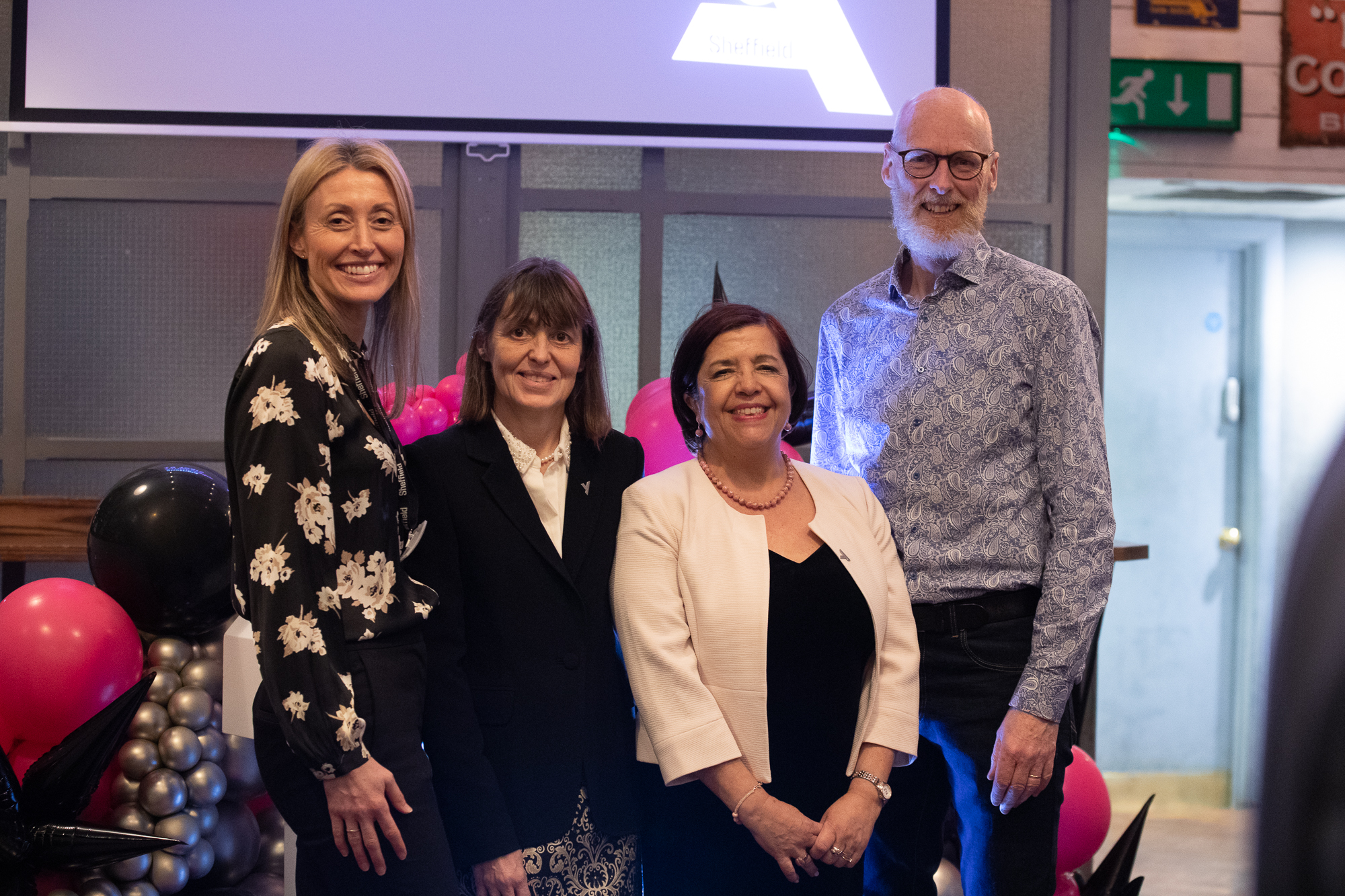 Four people standing together indoors at an event, positioned in front of a backdrop with a large screen displaying a partial logo. The group is surrounded by decorative balloon arrangements in black, pink, and silver. The setting includes grey paneled walls and visible signage in the background, suggesting a formal or celebratory occasion.