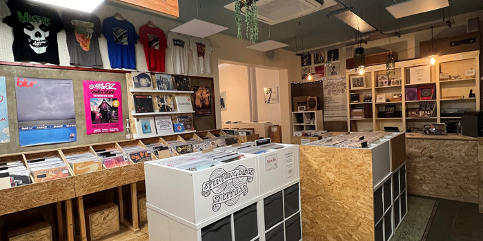 Interior of a record shop featuring wooden and white display units filled with vinyl records. A large sign overhead reads “NEW and REISSUE VINYL.” The walls display colorful posters and T-shirts, and the shop has a mix of wooden and terrazzo flooring. Hanging plants and ceiling lights add to the decor.