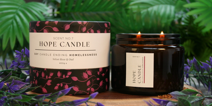 Hope Candle Scent No.7 in Velvet Rose & Oud fragrance, featuring a black and pink floral-patterned box and a lit amber glass jar candle with two wicks, displayed on a wooden base surrounded by green leaves and purple flowers.