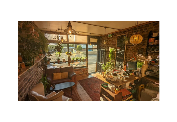 arm, sunlit café interior with exposed brick walls, wooden floors and large front windows. Hanging lights and numerous potted plants create a cosy atmosphere. A brown leather sofa, armchair, coffee table and patterned rug form a seating area near the window. To the right, a counter displays cakes, cups and a coffee machine, while golden evening light streams through the glass, illuminating the space.