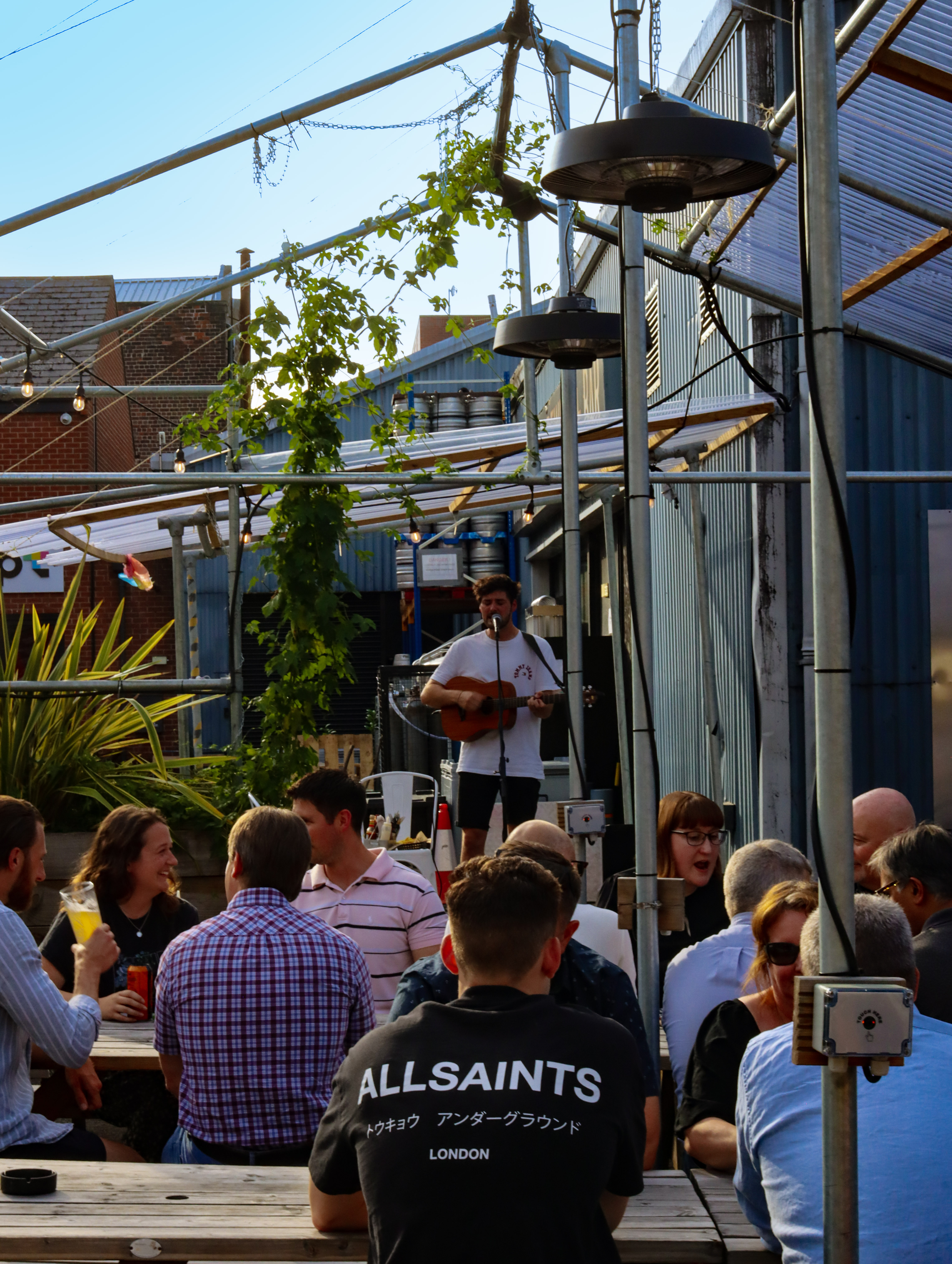 A singer performing with a guitar on a small outdoor stage, surrounded by metal frames and plants. People sit at picnic tables listening and drinking in a relaxed, sunny setting.