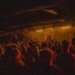 A packed audience inside a low‑lit venue faces a stage with bright amber lighting, where musicians perform. Raised hands and silhouettes fill the scene, creating a lively concert atmosphere.