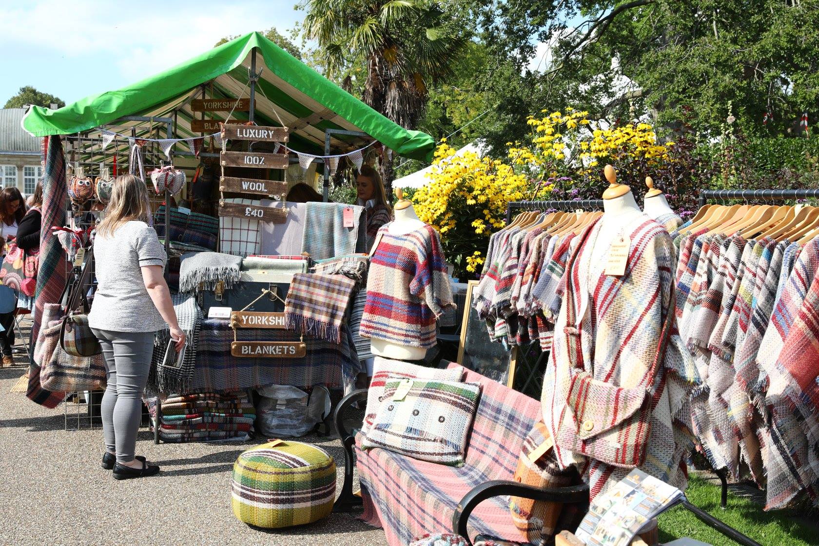  An outdoor market stall displays a variety of colourful wool blankets, throws, and ponchos arranged on racks, mannequins, and benches. A green canopy shades the stall, which also features cushions and a round pouffe in matching patterns. A wooden sign reads “Yorkshire Blankets” alongside smaller signs for luxury wool items. Bright yellow flowers and lush greenery surround the area, creating a vibrant and inviting setting.