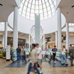 The interior of Meadowhall beneath a tall glass dome, showing escalators, white pillars, surrounding shops and shoppers moving through the space.