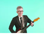 Studio portrait of a person wearing a black suit, white shirt and black tie, standing against a mint-green background while holding an electric guitar. The guitar has a dark red and black body with a light-coloured neck. The person is centred in the frame and wears round glasses, with a guitar strap over one shoulder.