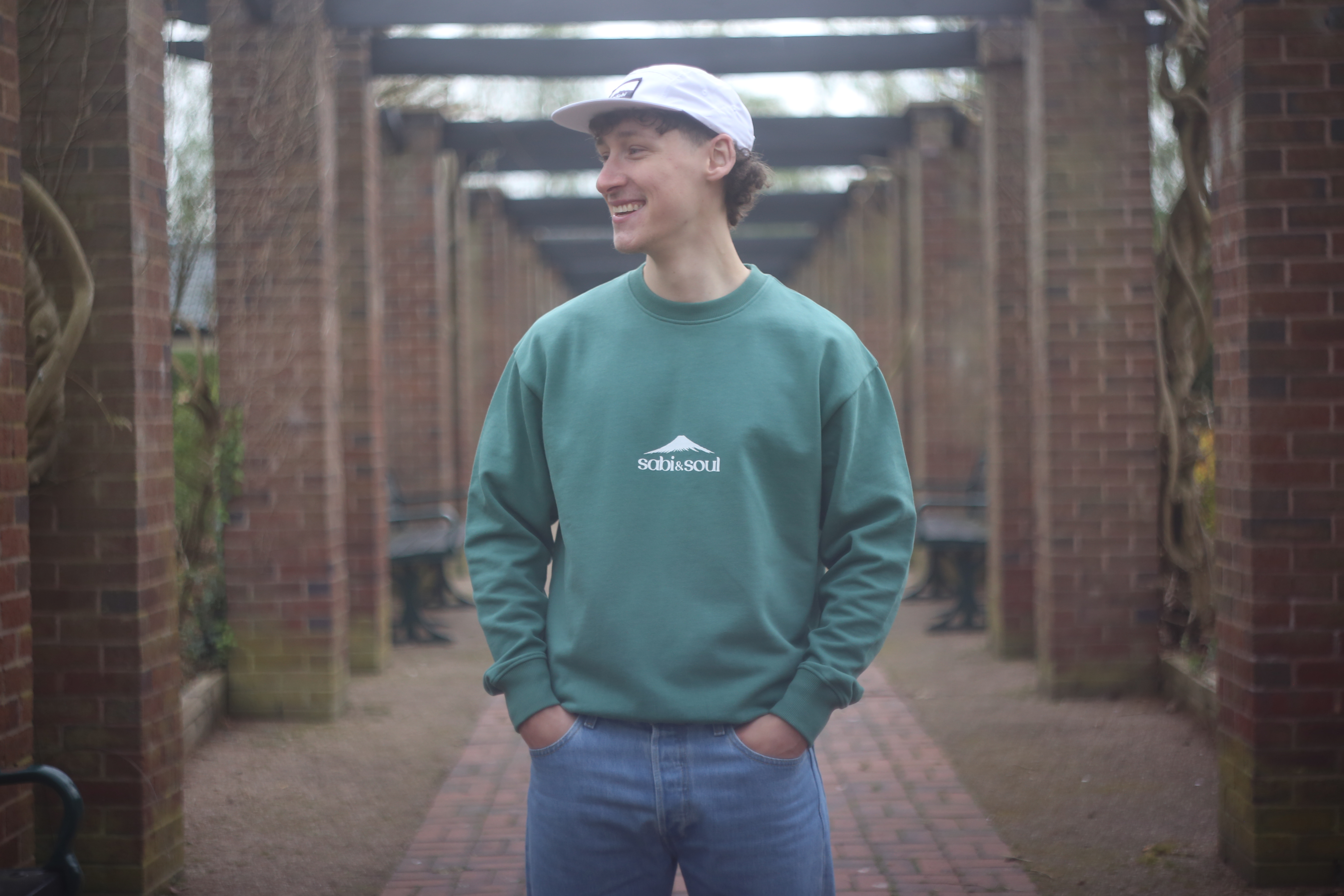 Person wearing a forest green sweatshirt with the “Sabi & Soul” logo printed in white on the chest, paired with light blue jeans and a white cap. The setting is an outdoor walkway framed by brick pillars and wooden beams.