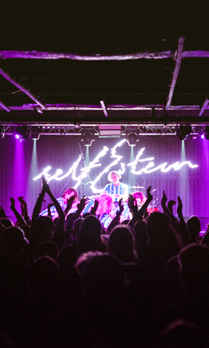 A crowded concert venue with an audience raising hands and cheering in front of a brightly lit stage. The stage is illuminated with vibrant purple lighting, and a performer stands at the center under a glowing neon-style sign that reads “self esteem.” The ceiling has exposed beams, and the atmosphere suggests an energetic live music event.
