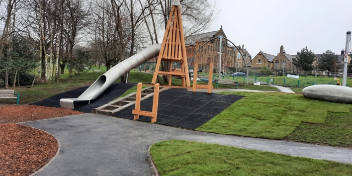 Playground with a large wooden-framed structure and a long, curved metal slide. The area is surrounded by grass, pathways, and leafless trees. Stone buildings and fencing are visible in the background under an overcast sky.