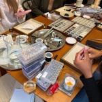 Jewellery-making workshop in progress around a large wooden table. Participants sit among trays of beads, tools, cords and jewellery findings, with one person threading materials by hand. Boxes of wooden and patterned beads, spools of cord, pliers and partially assembled items are spread across the workspace. A folder labelled “AKOEA” rests on a chair beside the table, creating a busy, hands-on craft studio atmosphere.