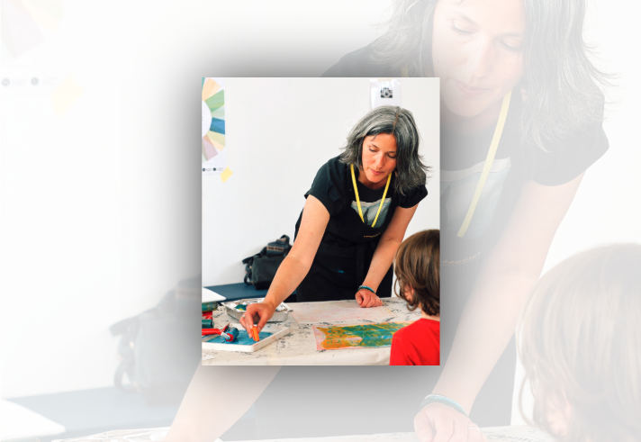 A workshop facilitator leans over a table demonstrating printmaking techniques to a child seated opposite. Art materials including rollers, ink trays and printed papers are spread across a covered work surface. The participant wears a yellow lanyard and reaches toward a roller coated with blue ink. The bright room has a plain white wall, with a colour wheel poster partly visible in the background.