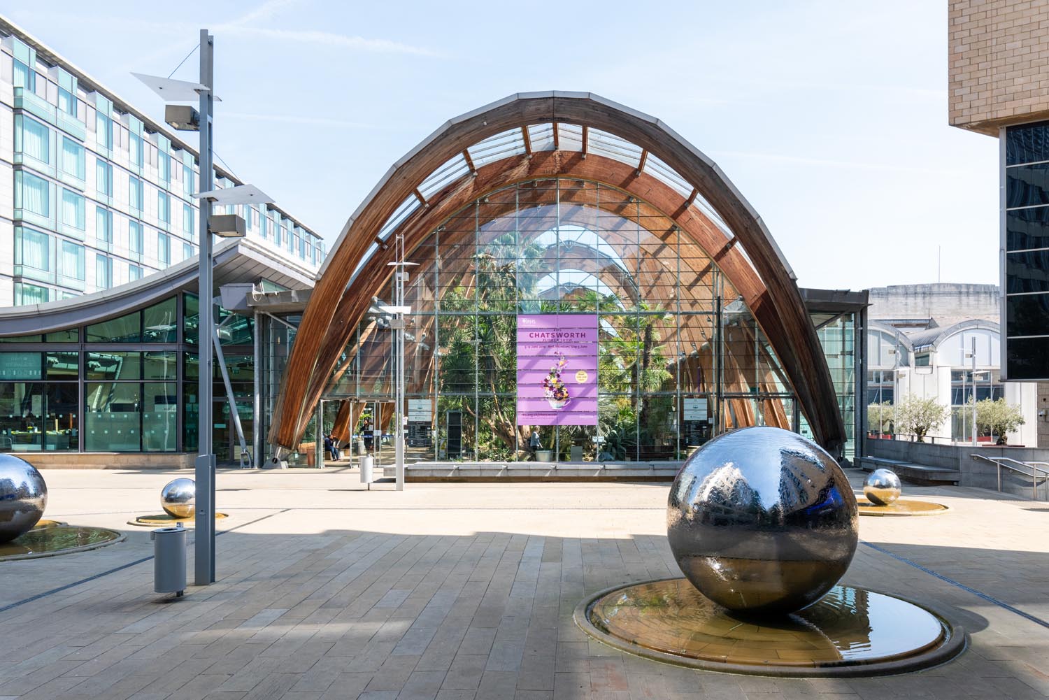 Exterior view of the Winter Garden in Sheffield, featuring a large arched glass and timber structure with a purple banner hanging inside. In the foreground, reflective silver spheres sit in shallow water features on a paved plaza, surrounded by modern buildings.