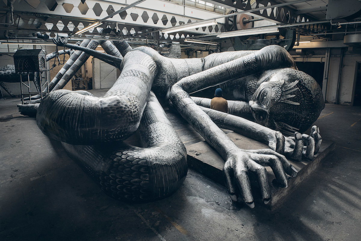 Large monochrome sculpture of a reclining humanoid figure with elongated limbs and intricate patterns, displayed inside an industrial-style warehouse with exposed beams and machinery.