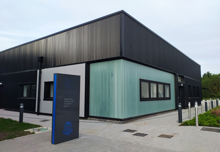 Modern single-story building with a mix of black metal cladding and light green panels, featuring large rectangular windows. A freestanding sign in front displays text, and the building is surrounded by a paved walkway with bollards and landscaped greenery.