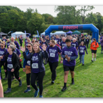 Participants wearing purple “Walk As One” event T-shirts walk across a grassy park after passing beneath a large blue inflatable arch marked “FINISH”. Several people wear numbered bibs, and event tents and flags are visible in the background. Trees surround the park, and a QR code, volunteers and spectators can be seen around the finish area, creating the atmosphere of a community fundraising walk.