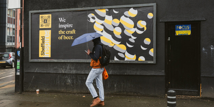 A person walking along a city street in the rain, holding an umbrella and passing a large billboard that reads “We inspire the craft of beer,” featuring bold graphic artwork on a dark wall.