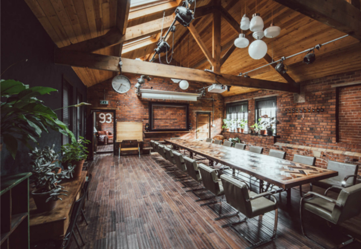 Inside The Chimney House . Spacious meeting room with exposed brick walls, wooden ceiling beams, and large skylights allowing natural light. A long rectangular table with modern chairs runs down the center of the room. Pendant lights and spotlights hang from the ceiling, and plants decorate the side tables and windowsills. A large wall clock and framed board are mounted on the far wall, adding to the rustic-industrial design.