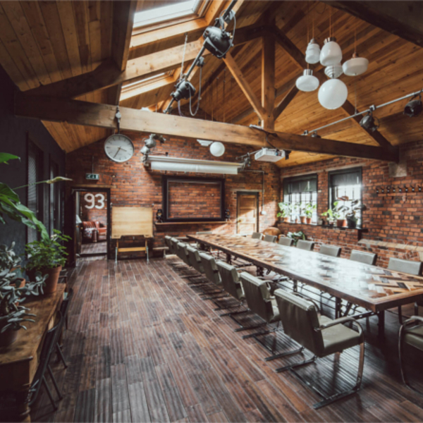 Inside The Chimney House . Spacious meeting room with exposed brick walls, wooden ceiling beams, and large skylights allowing natural light. A long rectangular table with modern chairs runs down the center of the room. Pendant lights and spotlights hang from the ceiling, and plants decorate the side tables and windowsills. A large wall clock and framed board are mounted on the far wall, adding to the rustic-industrial design.