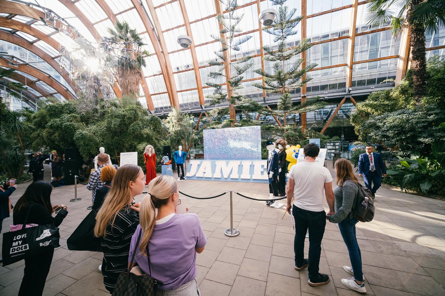A group of people gather inside a spacious, glass-roofed conservatory filled with lush greenery and tall trees. A large sign reading 'JAMIE' stands in the center. Some attendees take photos while others explore the surroundings, with natural light streaming through the glass ceiling.