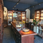 Interior of an art gallery with dark blue walls, wooden shelving and glass display cases. Sculptures, ceramics, jewellery and decorative objects are arranged throughout the space, with spotlights illuminating the exhibits. A long wooden display counter runs through the centre of the room, holding flowers and artwork, while stairs at the back lead to a raised area displaying additional pieces.