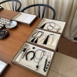 Display of handmade accessories arranged in shallow wooden trays on a wooden table. The collection includes black and white tassels, beaded bracelets, necklaces, keyrings and bag charms in monochrome tones. Several trays are branded “oicé.” The display sits beside black chairs, with cream curtains in the background and a woven natural-fibre rug below, creating a calm, minimalist craft market or showroom setting.