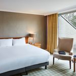 Bedroom with crisp white bedding and a armchair positioned in front of a large window allowing ample natural light.