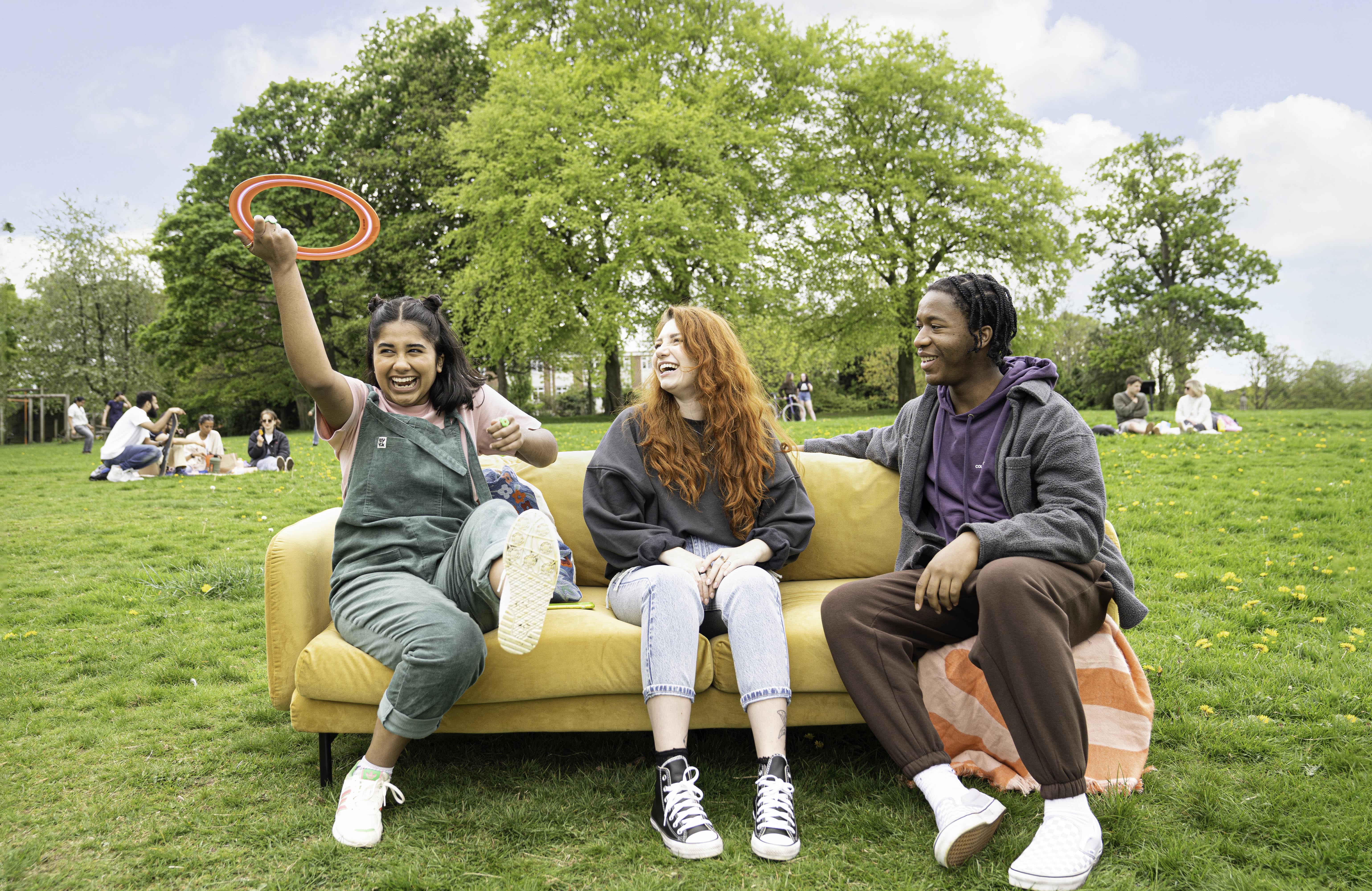 Three people sitting on a yellow sofa placed outdoors on a grassy field. One person on the left is holding up a red ring toy, while the others sit casually with legs crossed. In the background, there are groups of people relaxing on the grass and large green trees under a bright blue sky with scattered clouds.