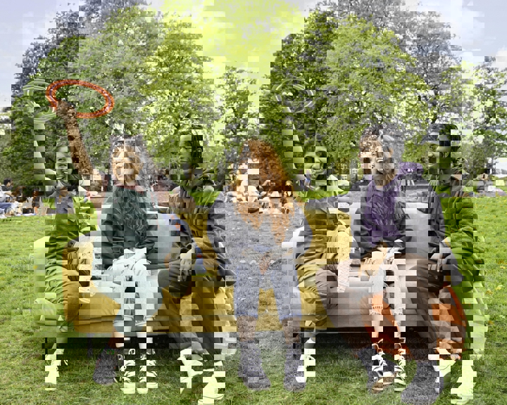 Three people sitting on a yellow sofa placed outdoors on a grassy field. One person on the left is holding up a red ring toy, while the others sit casually with legs crossed. In the background, there are groups of people relaxing on the grass and large green trees under a bright blue sky with scattered clouds.