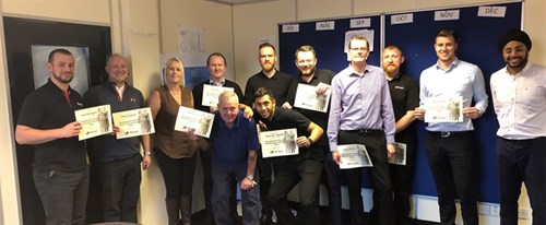 A dozen people standing in a workspace. They are all holding certificates and smiling at the camera.