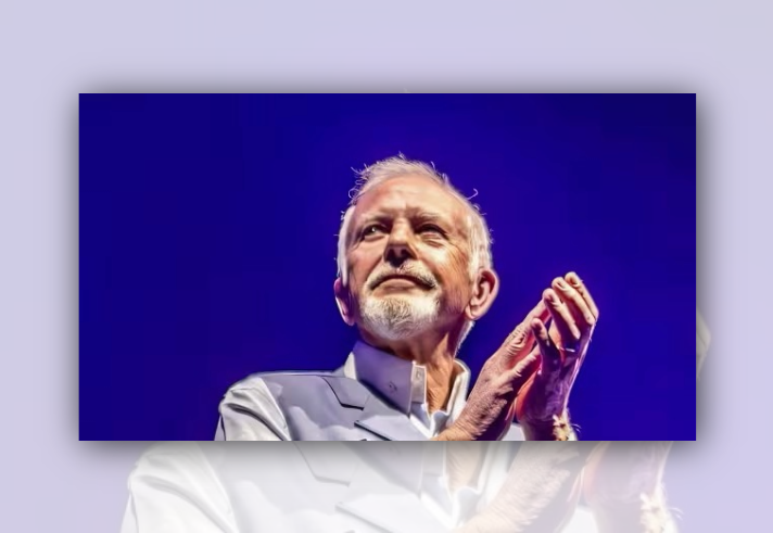Close-up stage photograph of a person with short white hair and a white beard wearing a light-coloured jacket. The person is shown from the chest up against a vivid blue background, with hands raised together near the face. Bright stage lighting highlights the face and clothing, creating a striking concert or public-event portrait.