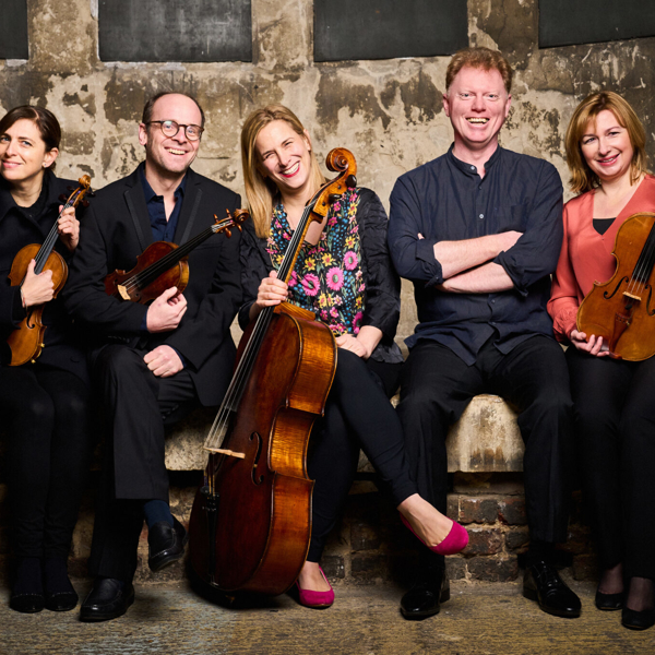 Five people (three women and two men) are sat on a low wall in a dilapidated building. One of the men and two of the women are holding violins and one of the women has a cello. 