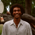 Photo of a person standing outdoors in front of a large fallen tree trunk in a park or woodland setting. Musical percussion instruments and woven shakers hang from and rest on the tree. The person wears a light-coloured shirt and necklace and stands centred in the image. Green grass and trees form a softly blurred background, creating a natural and relaxed atmosphere.