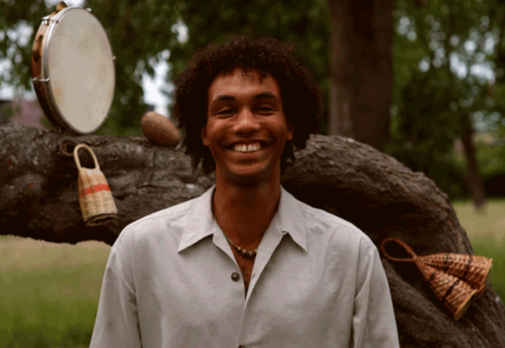 Photo of a person standing outdoors in front of a large fallen tree trunk in a park or woodland setting. Musical percussion instruments and woven shakers hang from and rest on the tree. The person wears a light-coloured shirt and necklace and stands centred in the image. Green grass and trees form a softly blurred background, creating a natural and relaxed atmosphere.