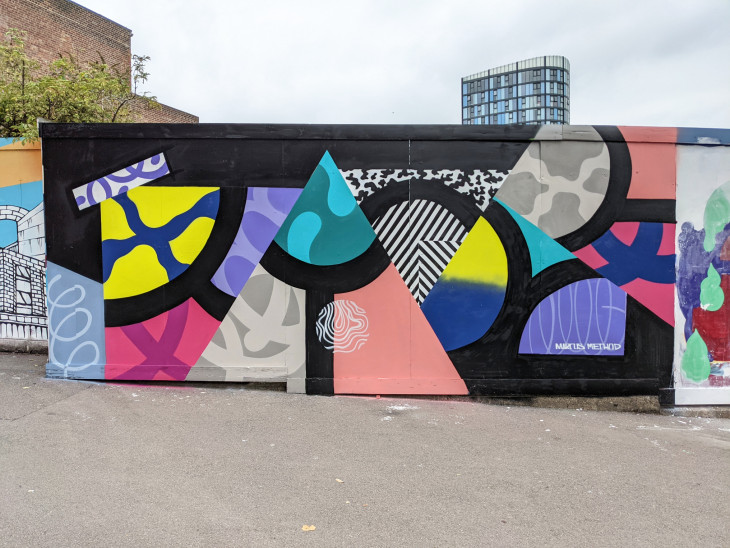 Street art mural featuring bold geometric shapes and patterns in vibrant colors including teal, pink, yellow, blue, and black. The design includes triangles, circles, and abstract textures on a black background, painted on an outdoor wall in an urban setting with a tall modern building visible in the background.