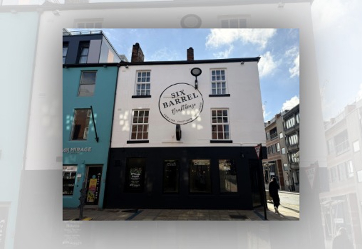 Street-level view of the Six Barrel Drafthouse, a white two-storey building with black trim and sash windows. A large circular sign on the front reads “Six Barrel Drafthouse” with “Cask Ales, Craft Beer, Fresh Coffee” around the edge. The ground floor has dark-painted shopfront windows displaying the words “Craft”, “Coffee” and “Cask”. A turquoise Mirage Vape Store is attached on the left, with modern city buildings and a Give Way sign visible on the right under a bright, partly cloudy sky.